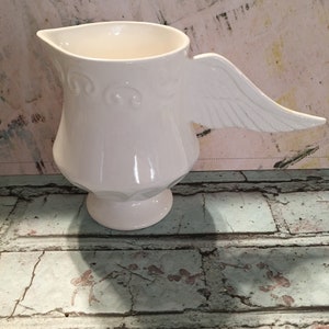 May include: White ceramic pitcher with a wing-shaped handle. The pitcher has a decorative design around the base.
