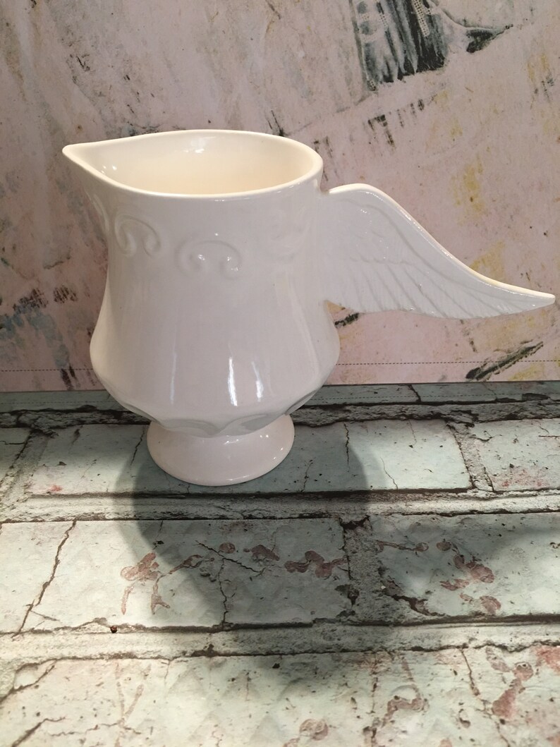 May include: White ceramic pitcher with a wing-shaped handle. The pitcher has a decorative design around the base.