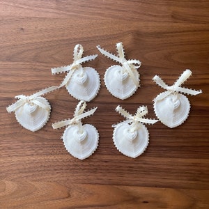 Puede incluir: Conjunto de seis adornos blancos en forma de corazón con una cinta de satén blanca y un borde de cuentas. Los adornos están dispuestos en círculo sobre una superficie de madera.