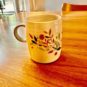 May include: A white ceramic mug with a floral design in shades of red, green, and yellow. The mug has a handle and is sitting on a wooden table.