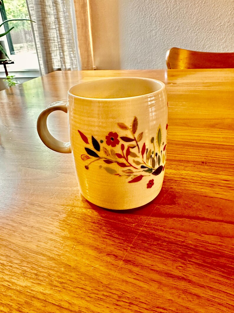 May include: A white ceramic mug with a floral design in shades of red, green, and yellow. The mug has a handle and is sitting on a wooden table.