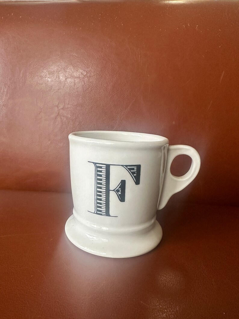 May include: White ceramic mug with a black letter F printed on the front. The mug has a wide base and a handle.