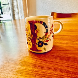 May include: A white ceramic mug with a floral design and the letter "P" in red, blue, and green. The mug is sitting on a wooden table.