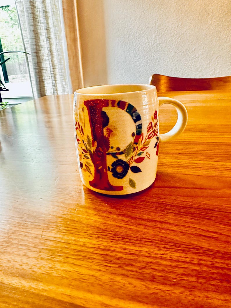 May include: A white ceramic mug with a floral design and the letter "P" in red, blue, and green. The mug is sitting on a wooden table.