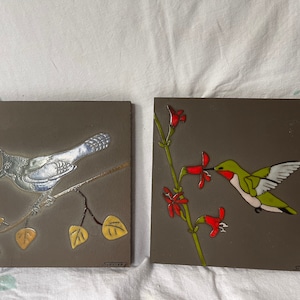 May include: Two ceramic tiles with painted bird designs. The first tile features a blue jay perched on a branch with yellow leaves. The second tile shows a hummingbird with green and red feathers, drinking nectar from a red flower.