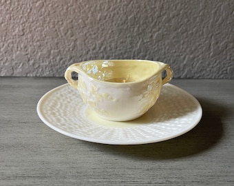 May include: A white ceramic creamer with a yellow glaze and a floral design. The creamer has two handles and is sitting on a white saucer with a textured design.