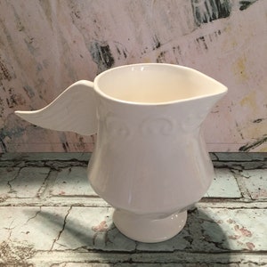 May include: White ceramic pitcher with a wing-shaped handle. The pitcher has a decorative design around the base.