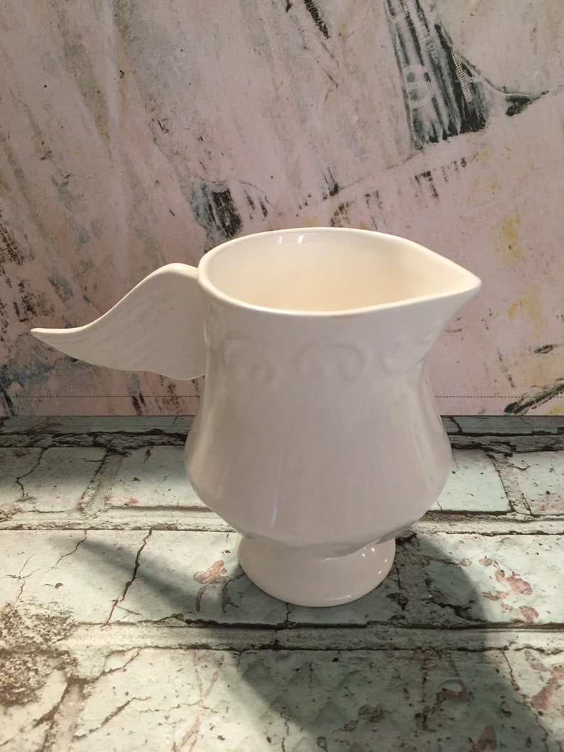 May include: White ceramic pitcher with a wing-shaped handle. The pitcher has a decorative design around the base.