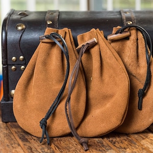 May include: Three brown suede pouches with black drawstrings and wooden toggles are displayed in front of a dark brown wooden chest with brass accents. The pouches are on a wooden surface.