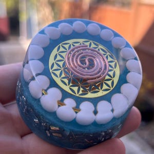 Op de afbeelding: Een blauw-witte orgone-piramide met een koperen spiraal en een gouden geometrisch ontwerp. De piramide is rond met witte en gouden accenten. Het midden heeft een koperen spiraal over een gouden geometrisch patroon.