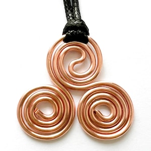 Puede incluir: Un collar con un colgante de alambre de cobre de color cobre con un diseño de triple espiral. El collar está en un cordón negro.