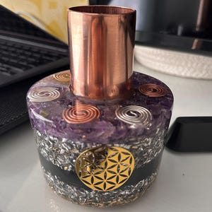 May include: An orgone energy generator with a copper cylinder on top. The base is a mix of purple amethyst, silver metal, and a gold-coloured geometric design. The generator is on a white surface.