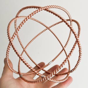 May include: A copper-toned, spherical sculpture made from interwoven, twisted metal rods. The open design creates a geometric pattern, with the rods intersecting to form a three-dimensional orb. The sculpture is held in a hand against a white backdrop.