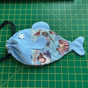 May include: A blue fabric drawstring bag shaped like a fish with a white star button. The bag is decorated with a winter scene featuring snowmen and robins.