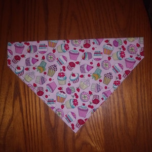 May include: Pink fabric dog bandana with a pattern of colourful cupcakes and cherries.
