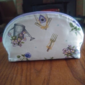 May include: A white zippered pouch with a lavender zipper. The pouch has a floral print with a watering can, a birdhouse, a garden fork, and blue flowers.