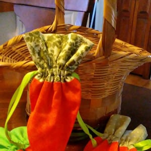May include: Two orange carrot-shaped fabric bags with green and white floral fabric tops. The bags are sitting in a wicker basket.