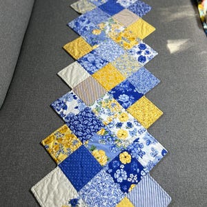 Puede incluir: Camino de mesa acolchado con diseño de patchwork. El camino de mesa está hecho de piezas de tela en forma de diamante en tonos azules, amarillos y blancos. Se ven estampados florales y de lunares. El camino de mesa mide unos 152 cm de largo.