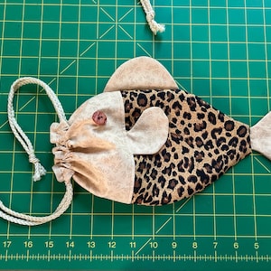 May include: A fish-shaped drawstring bag, handmade from fabric. The head and tail are tan with a floral pattern, while the body has a leopard print. A wooden button secures the drawstring. The bag is approximately 25 cm long.