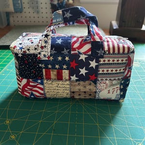 May include: A patchwork tote bag with a handle, featuring a patriotic design with red, white, and blue stars and stripes. The bag has a rectangular shape and is on a green cutting mat.