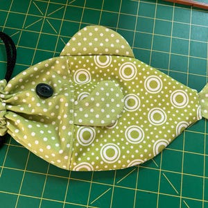 May include: A fish-shaped fabric pouch in light green with a white polka dot and concentric circle pattern. The pouch features a black button eye, a gathered opening at the head, and black cord drawstrings attached to the side for closure.