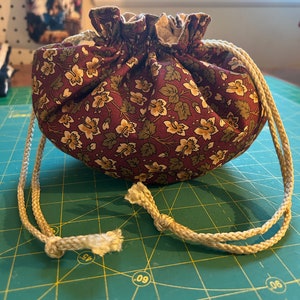 May include: A small drawstring bag made of burgundy fabric with a floral pattern. The bag has a cream-coloured drawstring and is sitting on a green cutting mat.