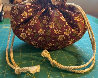 Pode incluir: Uma pequena bolsa de cordão feita de tecido bordô com um padrão floral. A bolsa tem um cordão de cor creme e está sentada em um tapete de corte verde.