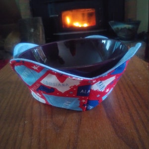 May include: A red, white and blue fabric bowl cosy with state outlines and stars. The cosy is on a wooden surface and is holding a dark purple bowl.
