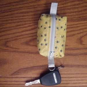 May include: A small yellow zippered pouch with a white zipper and a white strap. The pouch is covered in a pattern of small black bees. A black and silver key is attached to the strap.