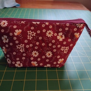 May include: A burgundy zippered pouch with a white floral pattern. The pouch is made of fabric and has a zipper closure.