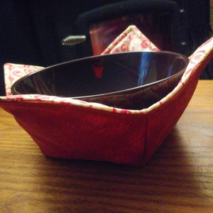 May include: A red fabric bowl cosy with a white and red floral pattern. The cosy is holding a dark brown bowl.