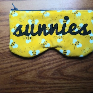 May include: A yellow fabric zippered pouch with a black embroidered design of bees and the word "sunnies".