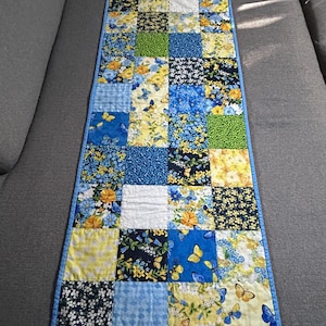 May include: A rectangular quilted table runner with a patchwork design. The squares feature floral and butterfly patterns in blue, yellow, and white, with a blue border. The runner measures approximately 30 cm wide and 76 cm long.