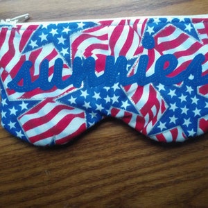 May include: A red, white and blue fabric zippered pouch with the word "sunnies" embroidered in blue. The fabric features a pattern of stars and stripes.