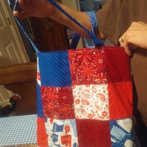 May include: A red, white and blue patchwork tote bag with a variety of patterns, including stars, paisley and a map of the United States. The bag has two blue straps.