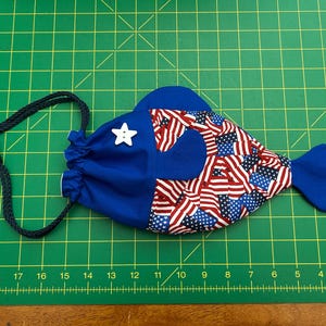 May include: A fish-shaped fabric drawstring pouch featuring a solid blue head section with a white star-shaped button and a body patterned with red, white, and blue American flag motifs. The pouch includes a dark blue braided cord closure.