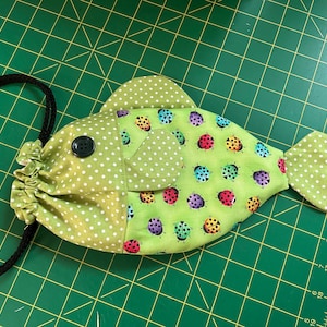 May include: A fish-shaped fabric pouch featuring a green and white polka dot pattern on the head and tail, and a light green body printed with colourful ladybirds. The pouch includes a black button eye and a black drawstring cord for closure.