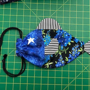 May include: A fabric fish-shaped drawstring bag. It features a blue and white star pattern, blue floral print, and black and white striped details. The bag has a black drawstring and is on a green grid surface.