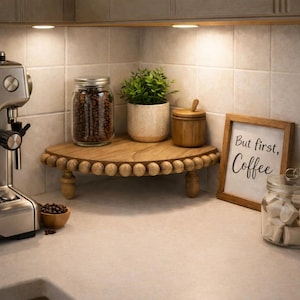 Könnte beinhalten: Ein hölzernes Eckregal mit Perlenrand, auf dem ein Glas mit Kaffeebohnen, eine kleine Topfpflanze und ein Holzbehälter stehen. Daneben befindet sich ein gerahmtes Schild mit dem Text "But first, Coffee" sowie ein Glas mit weißen Zuckerwürfeln.