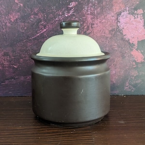 May include: A brown ceramic jar with a white lid. The jar has a rounded shape and a smooth surface. The lid has a small knob on top.