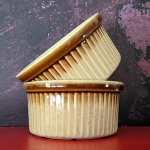 May include: Two brown and beige ceramic ramekins with a ribbed design. The ramekins are stacked on top of each other.