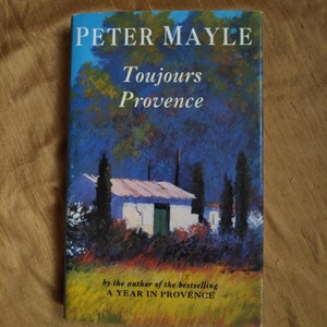May include: A book cover with a blue and yellow painted landscape featuring a white house with a green door. The title of the book is "Toujours Provence" by Peter Mayle. The text "by the author of the bestselling A YEAR IN PROVENCE" is also on the cover.