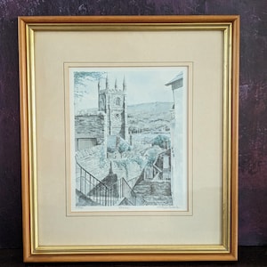 May include: A framed black and white print of a church with a tall steeple. The print is in a wooden frame with a gold trim. The print is titled "Penryn" and is signed by the artist.