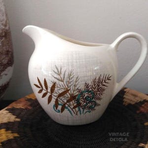 May include: White ceramic creamer with a brown and teal leaf design. The creamer has a textured surface and a rounded body.