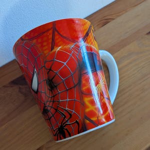 May include: A ceramic mug featuring a vibrant red and orange graphic print of the character Spider-Man. The design includes a detailed black web pattern covering the surface, a white eye mask detail, and a small black spider icon near the base.