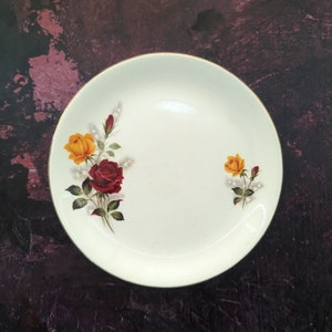 Könnte beinhalten: Ein weißer Keramikteller mit goldfarbenem Rand zeigt ein Blumenmuster mit roten und gelben Rosen. Der Teller ist perfekt zum Servieren von Desserts oder Vorspeisen.