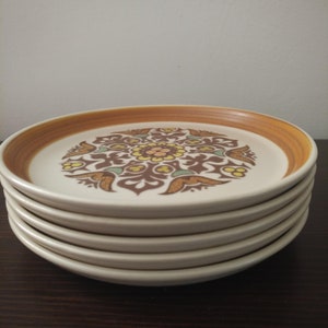 May include: A stack of six ceramic plates. Each plate has a cream-coloured centre with a brown, yellow, and green floral design. The rims are a warm brown colour. The plates are stacked on top of each other.
