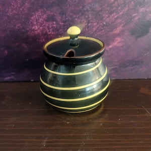 May include: A black and yellow striped ceramic honey pot with a lid and a small knob. The pot has a round shape and a small opening for a spoon. The lid is black with a yellow knob.