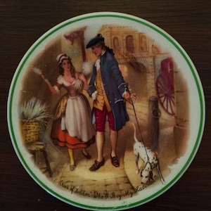 May include: A vintage ceramic plate with a green border depicts a man and woman in 18th-century clothing walking down a cobblestone street. The man is wearing a blue coat and the woman is wearing a white dress with a red and orange skirt. A dog is walking behind them. The text on the plate reads "Cries of London - Who'll Buy My Lavender."