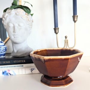 May include: A glossy, octagonal ceramic bowl featuring a deep brown glaze with lighter cream-colored drip accents along the top rim. The bowl is supported by a matching octagonal pedestal base, showcasing a smooth, reflective finish throughout.
